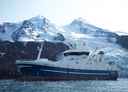 Blå og hvis fiskebåt foran øy med snøkledde fjell og en isbre.