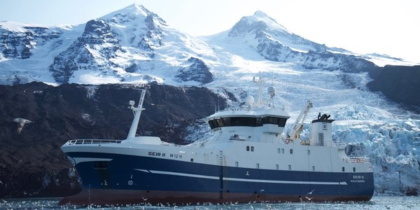 

Blå og hvis fiskebåt foran øy med snøkledde fjell og en isbre.