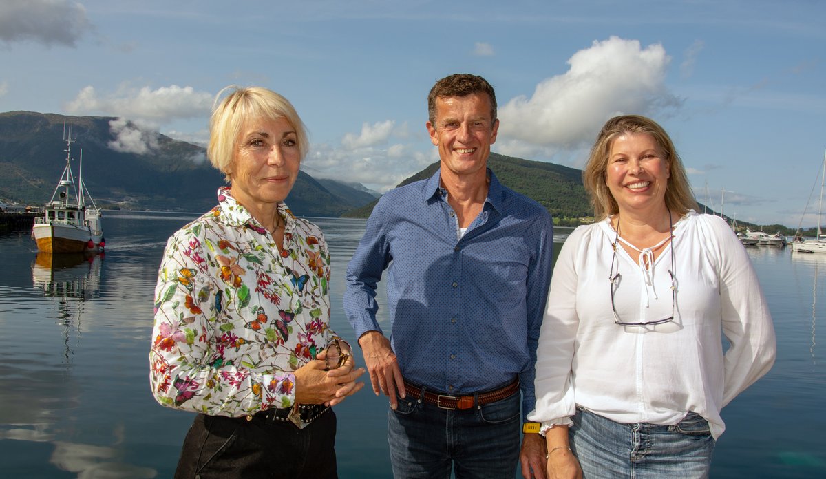 
To kvinner og en mann står og smiler i sola, med fjord, fjell og en fiskebåt i bakgrunnen.