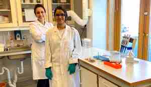 To kvinner i kvite labfrakkar på eit laboratorium, dei smiler mot kamera.