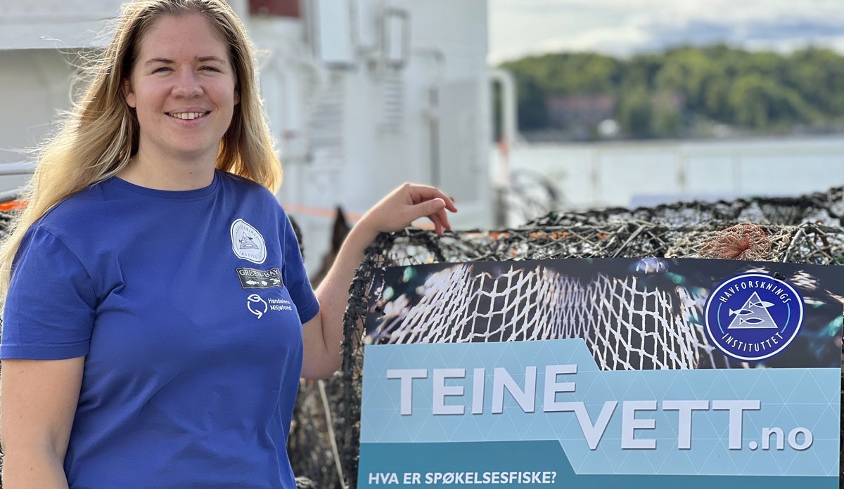 
Kvinne smiler til kamera, står foran plakat
