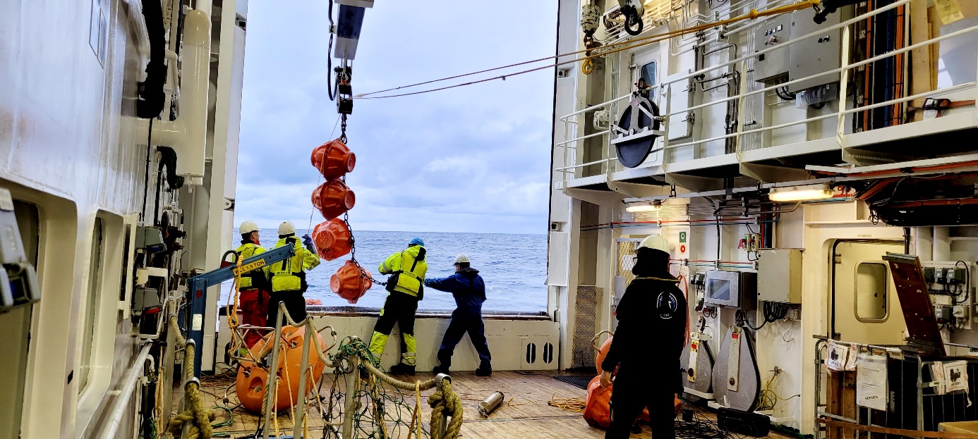 Bildet viser at riggene blir heist obord ved hjelp av kran gjennom lasteåpningen. Det er fire fra mannskapet som holder på med dette, og en person i bakgrunnen. 