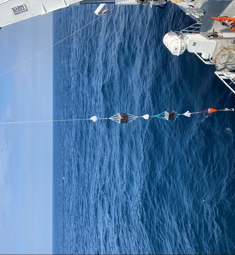 Picture showing a rope hanging from the RV’s winch. On the rope there is three sets of white bags holding zooplankton, in-between the 3 bags there are 2 black bokes (zooplankton camera cage) at the end of the rope there is a weight. Above the weight there is a drift anchor.