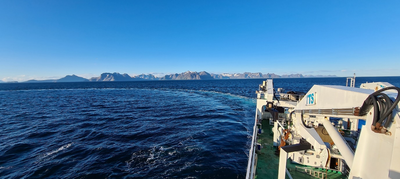 Photo taken by Ivar Wangen taken from high on the ship lookin bac over the rear and side of the vessel in bright sunlight. The sky is blue, the sea is calm. It is clear that the ship has been turning a circle from its wake, and the Troms coastal mountains are visible in the background with patches of snow on top.