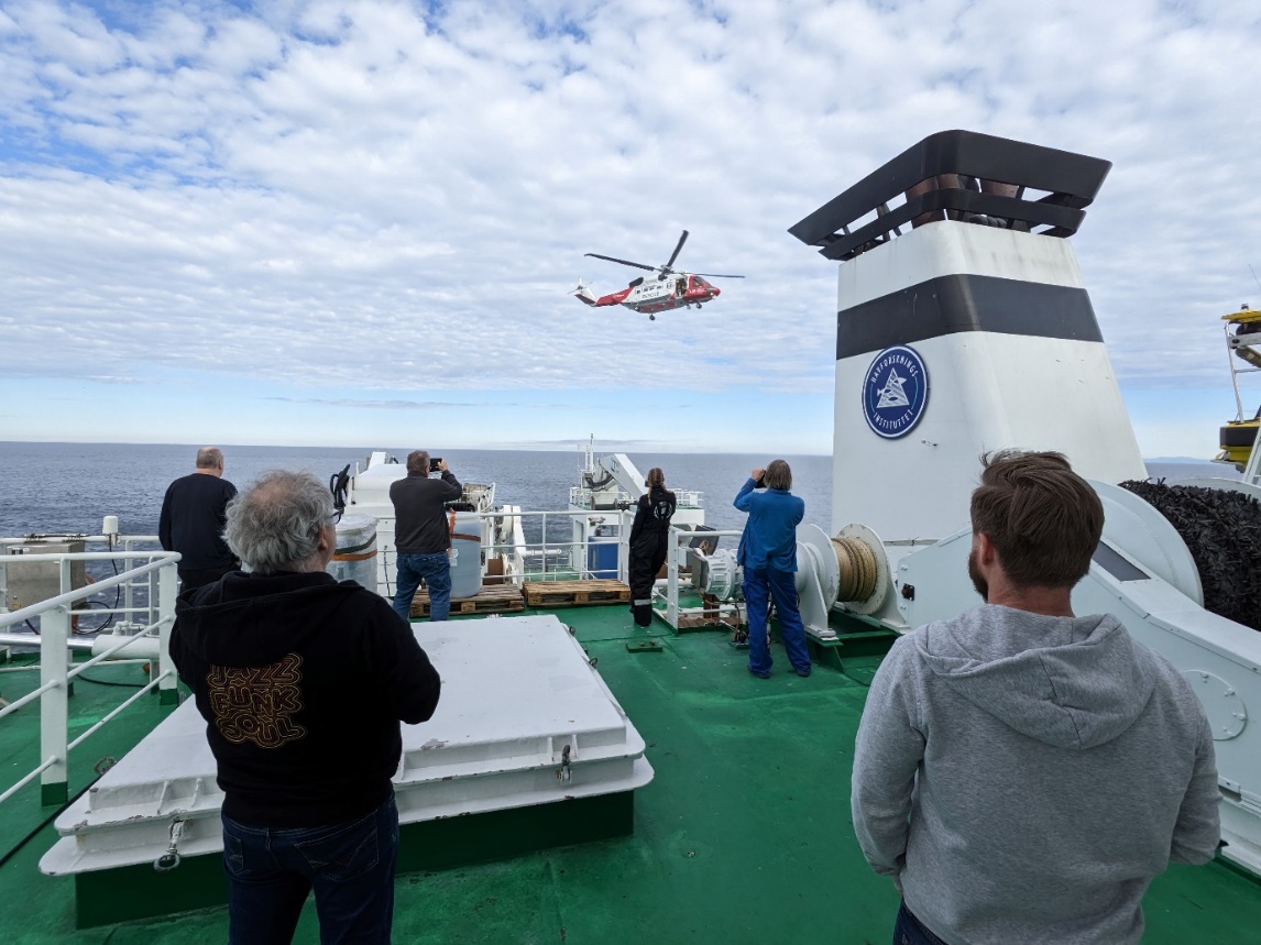 Photo of people out on an upper deck of the ship with their backs to the camera as they watch the search and rescue helicoper hover over the rear deck ready to do som practices for 20mins