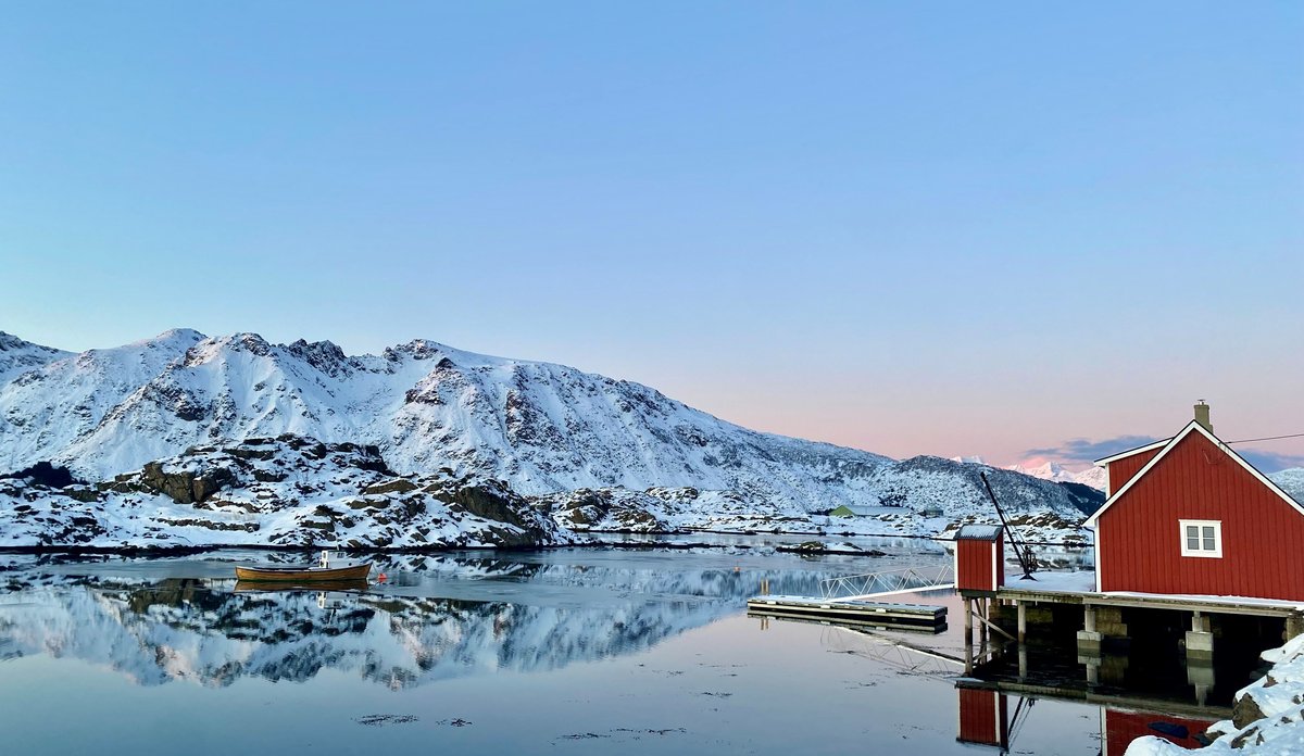 
Rødt naust ved havet, båt ligger på blikkstille vann med lave fjell i bakgrunnen. 