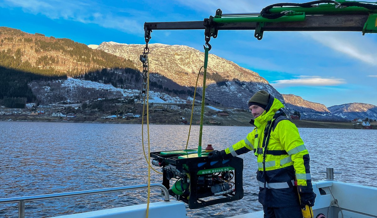 En mann med kjeledress står på dekk av en båt. Han er i ferd med å senke en ROV ned i fjorden.