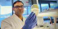 

En forsker i hvit labfrakk og blå plasthansker er på labben. I hånden holder han en petriskål med prøver.