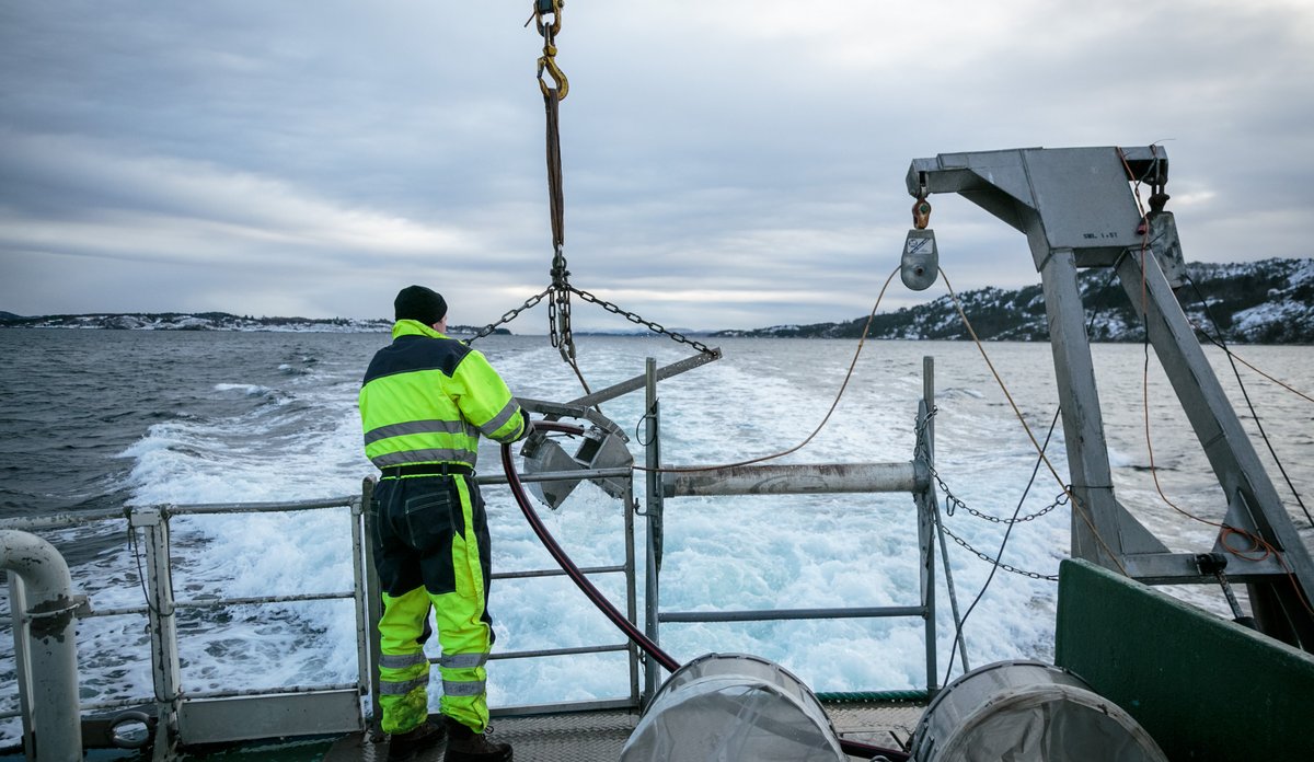 
Mann i kjeledress på dekk av et forskningsfartøy i fart, med grabb og trål.