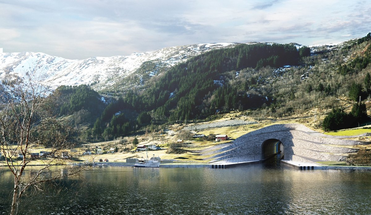 
tunnel i fjorden som går inn i en grønn bakke, med snøkledde fjell i bakgrunnen 